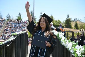 A graduate from Moorpark College wearing a cap and gown with a black stole waving at her family from the stage after accepting her diploma