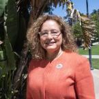 A woman with curly hair, wearing glasses and an orange blazer, smiles outdoors in a sunny park setting. She has a "Ventura College" button pinned to her jacket, with lush green foliage and a clear blue sky in the background.