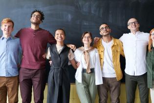 6 teachers of different ethnicities and genders standing with their arms linked and smiling in front of a blackboard 