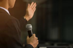 Photo of a person giving a speech while holding a microphone in one hand and gesturing with the other.