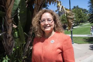 A woman with curly hair, wearing glasses and an orange blazer, smiles outdoors in a sunny park setting. She has a "Ventura College" button pinned to her jacket, with lush green foliage and a clear blue sky in the background.